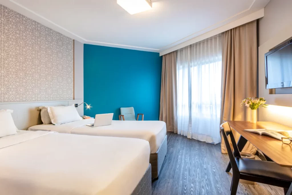 Um quarto de hotel com duas camas de solteiro brancas no Mercure Florianópolis, mostrando um notebook em cima de uma cama, uma parede azul e papel de parede com estampa geométrica, e uma área de trabalho com mesa e televisão.