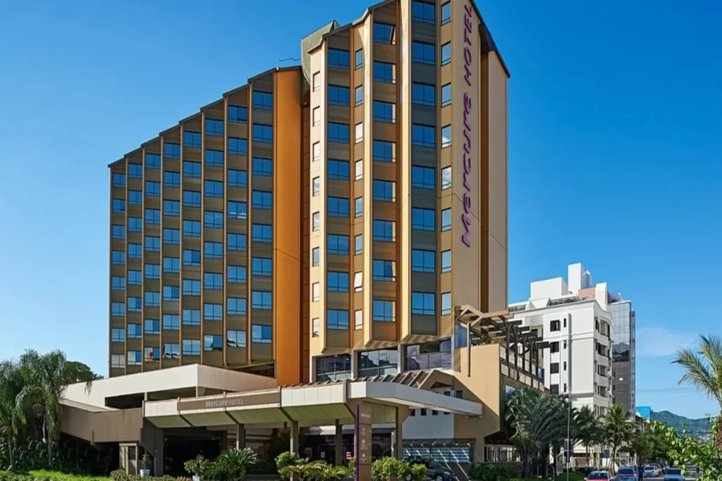 A fachada externa do hotel Mercure Florianópolis, um edifício em forma de 'V' com janelas azuis, o nome do hotel em roxo na lateral e a entrada coberta, sob um céu azul.