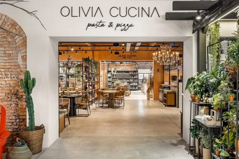 Entrada do restaurante Olivia Cucina, com o letreiro "pasta & pizza" acima, mostrando o ambiente interno com uma grande variedade de vinhos e produtos, uma área com mesas e cadeiras, e decorações com plantas e tijolos.