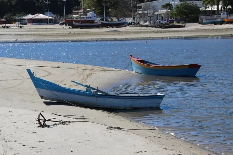 Dois barcos de pesca, um azul e um colorido, na areia e na água da praia de Ponta das Canas em Florianópolis, com a vila de pescadores e barcos maiores atracados ao fundo.