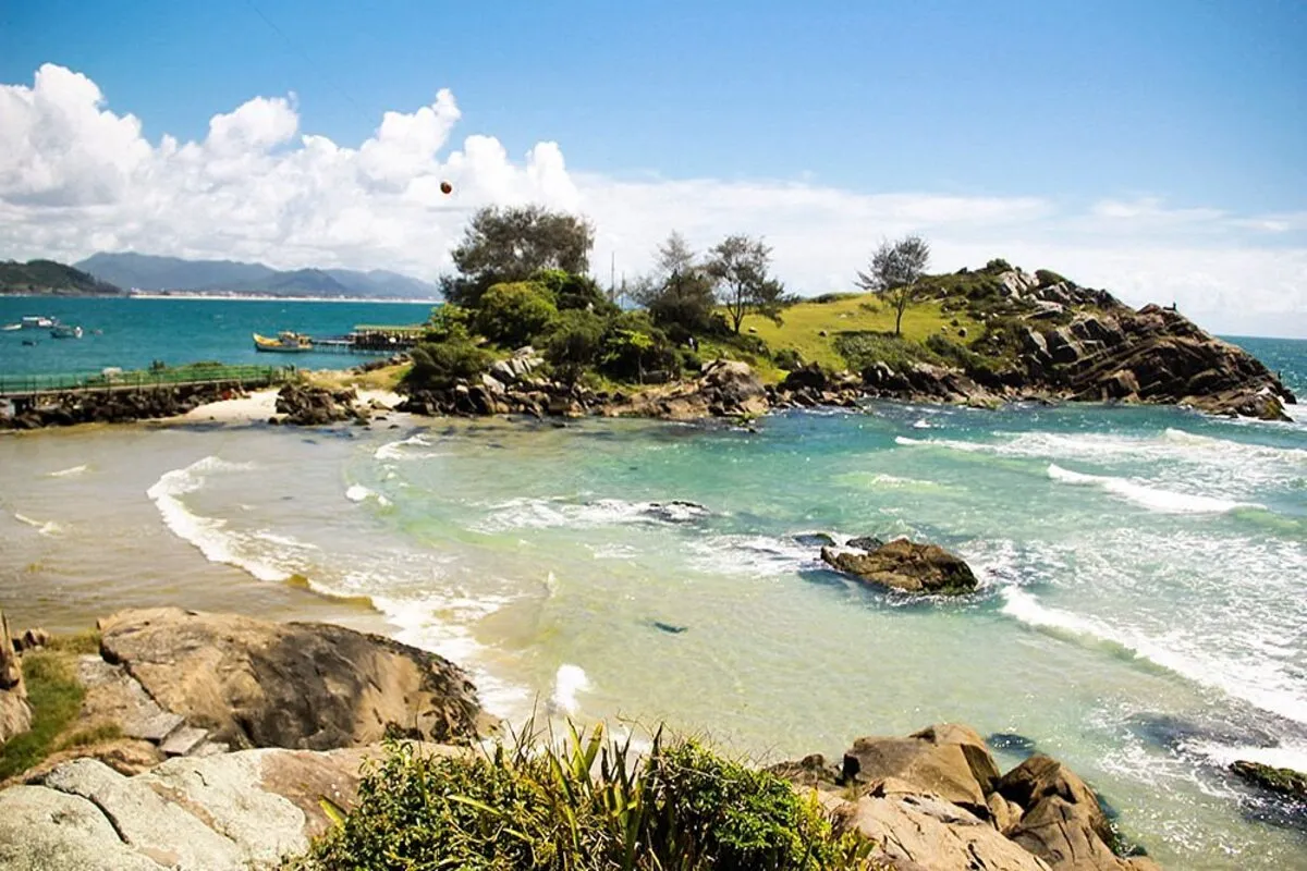 Vista da Praia do Matadeiro em Florianópolis, com mar azul cristalino, ondas leves e formações rochosas cercadas por vegetação.