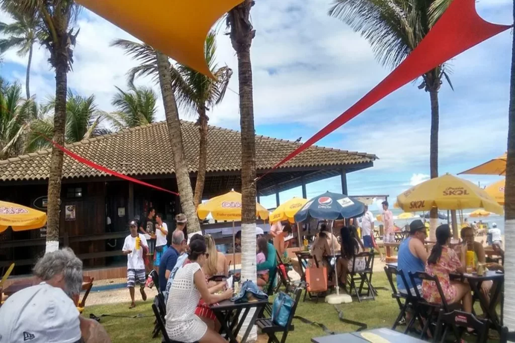 Vista de um restaurante à beira da praia em Florianópolis, o Timoneiro, com várias mesas e cadeiras de madeira em um gramado. Guarda-sóis amarelos com o logo "Skol" e palmeiras decoram o ambiente, e as pessoas estão sentadas, desfrutando de uma refeição.