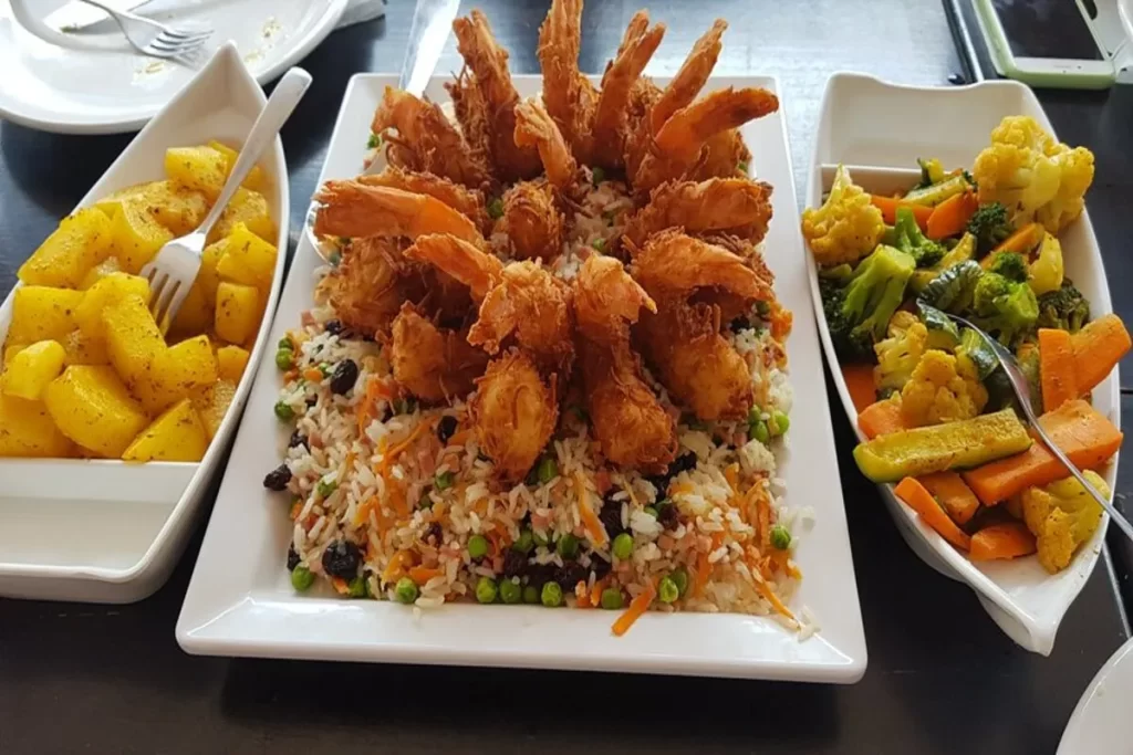 Um prato completo e farto de camarão empanado frito, servido sobre uma base de arroz com legumes e uva-passa. O prato é acompanhado por duas travessas laterais, uma com batatas salteadas e a outra com legumes cozidos no vapor.