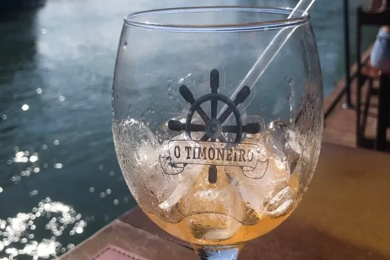 Um copo de drink de vidro transparente com gelo e o logo "O Timoneiro" (um timão de navio) em preto. O copo está sobre uma mesa, com o mar e o reflexo do sol ao fundo, sugerindo um ambiente à beira-mar.