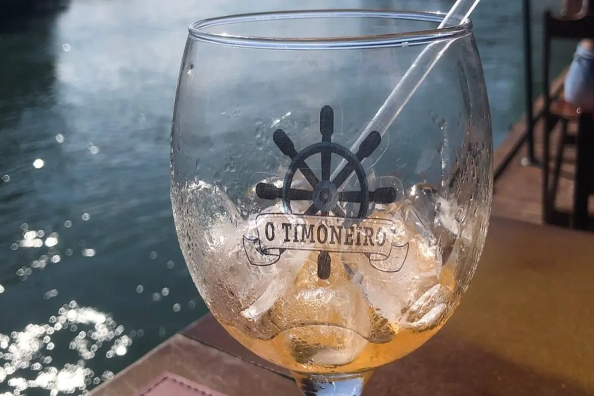 Um copo de drink de vidro transparente com gelo e o logo "O Timoneiro" (um timão de navio) em preto. O copo está sobre uma mesa, com o mar e o reflexo do sol ao fundo, sugerindo um ambiente à beira-mar.
