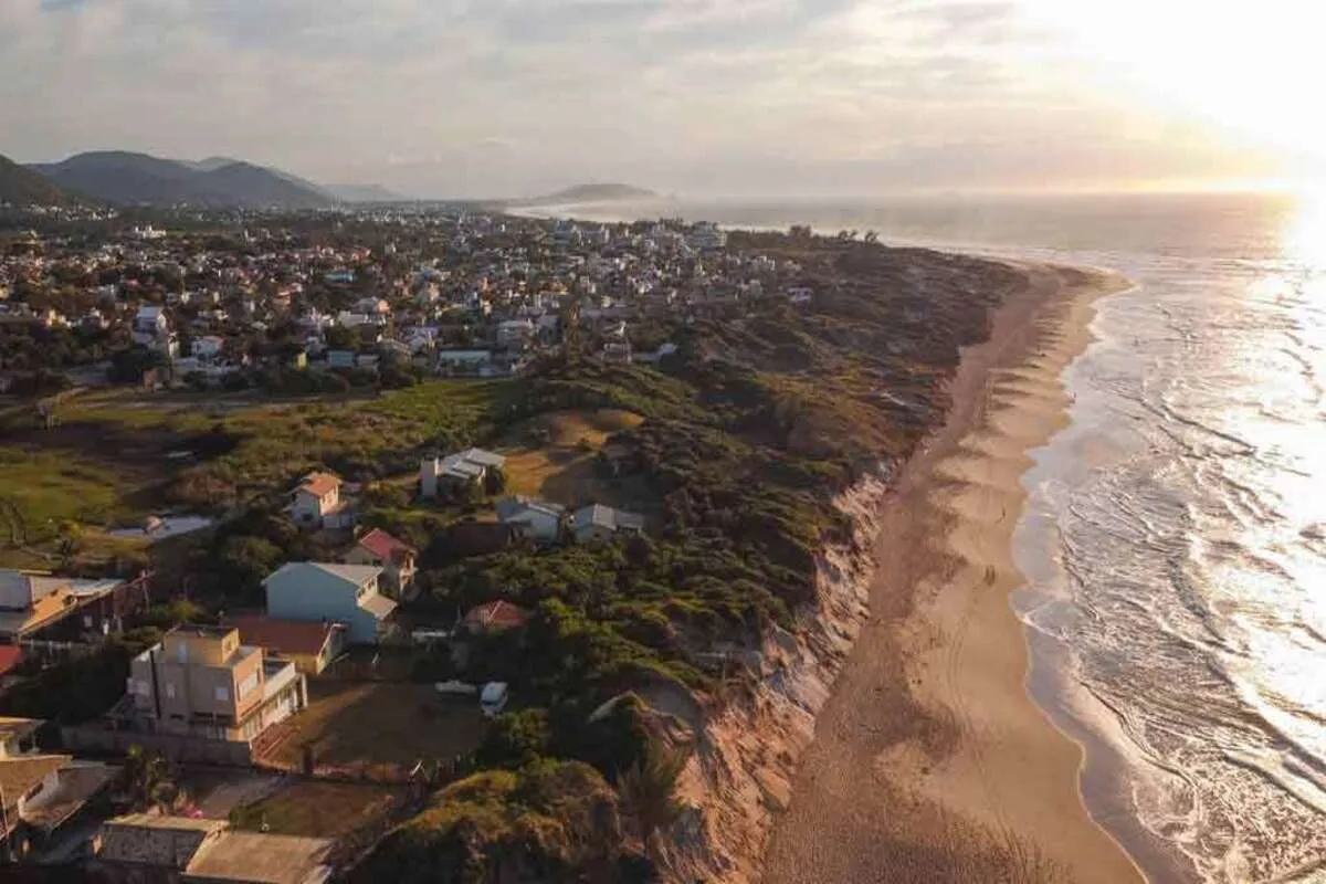 Vista aérea de uma paisagem costeira com uma longa faixa de areia dourada e a orla marítima com ondas. Há dunas e vegetação densa separando a praia de uma área residencial com casas de diferentes tamanhos e estilos. O sol está baixo no horizonte, refletindo uma luz brilhante e dourada sobre o oceano.