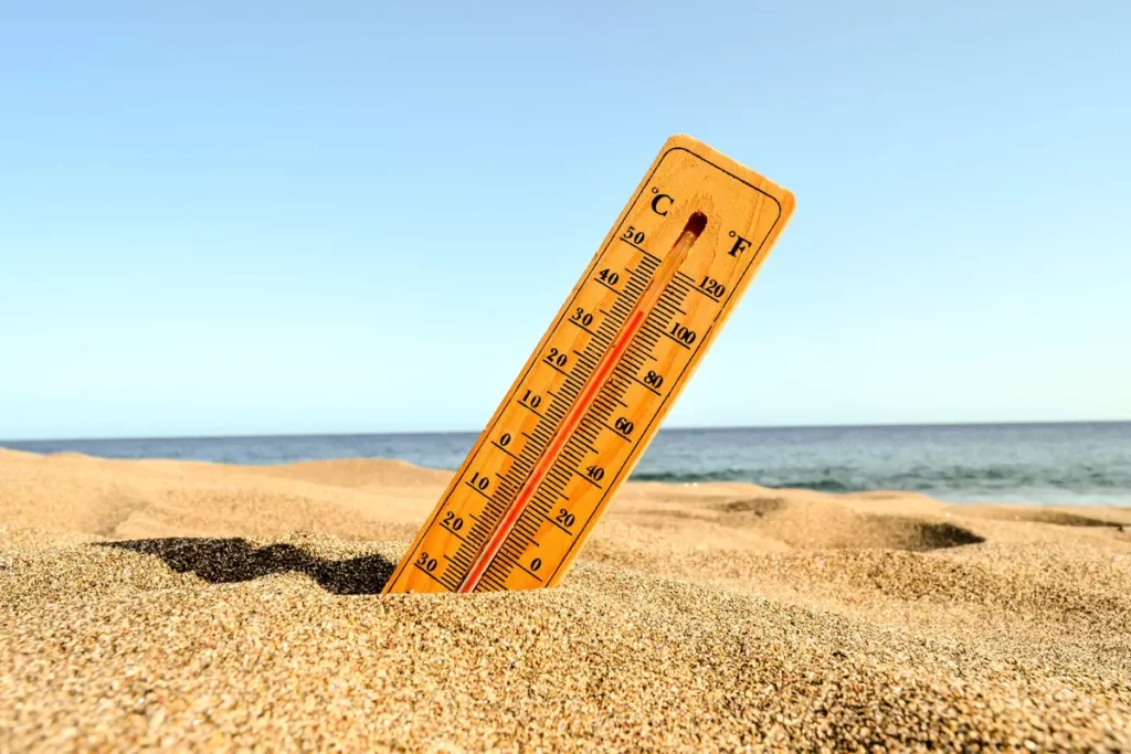 Um termômetro de madeira, com escala em graus Celsius e Fahrenheit, está fincado em uma areia de praia com reflexos dourados. A coluna de mercúrio está subindo, indicando uma temperatura alta, enquanto o fundo mostra o oceano e um céu azul claro.