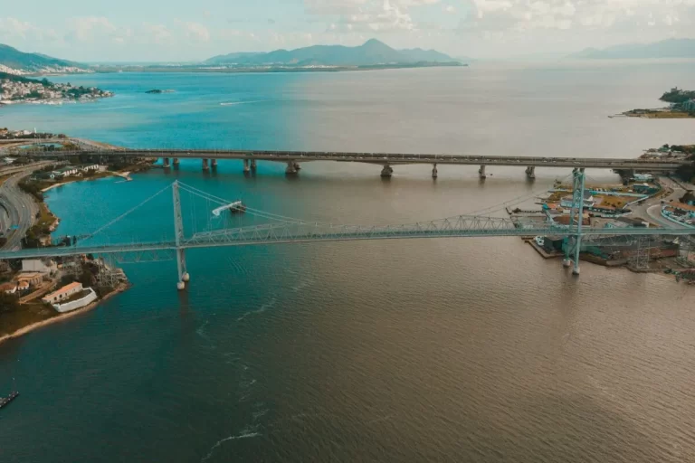 Vista aérea da Baía Sul em Florianópolis, mostrando a Ponte Hercílio Luz em primeiro plano e, ao fundo, a Ponte Colombo Salles. As pontes se estendem sobre a água, conectando a ilha de Florianópolis ao continente. A imagem também mostra a vegetação costeira, o céu com nuvens esparsas e o contraste de cores entre o oceano em tons de azul e marrom.