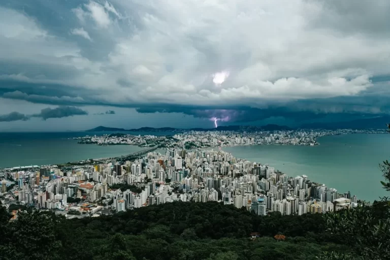 Vista aérea panorâmica de Florianópolis em um dia de tempestade, com nuvens de chuva escuras sobre a paisagem urbana. Um raio ilumina o céu no horizonte, com a cidade e as pontes entre a ilha e o continente visíveis.