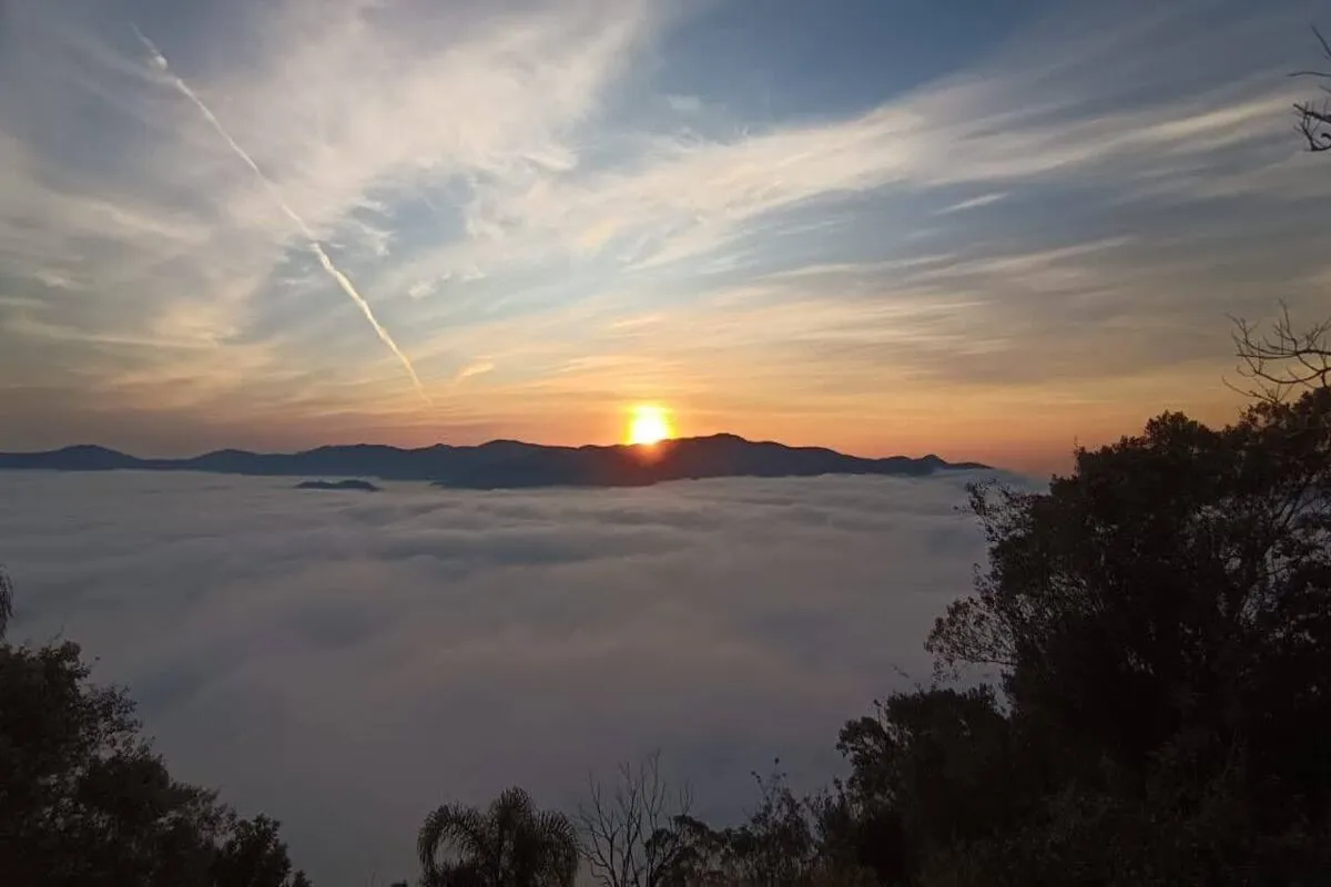 Vista panorâmica de um vasto mar de neblina cobrindo um vale ou cidade ao amanhecer, com o sol nascendo e iluminando o horizonte. As silhuetas de uma cordilheira de montanhas e de árvores no primeiro plano se destacam contra o céu colorido.