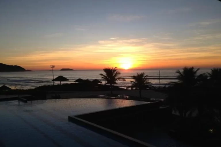 Pôr do sol na praia com o sol se pondo no horizonte, palmeiras e quiosques à beira-mar, criando um cenário tropical e relaxante.