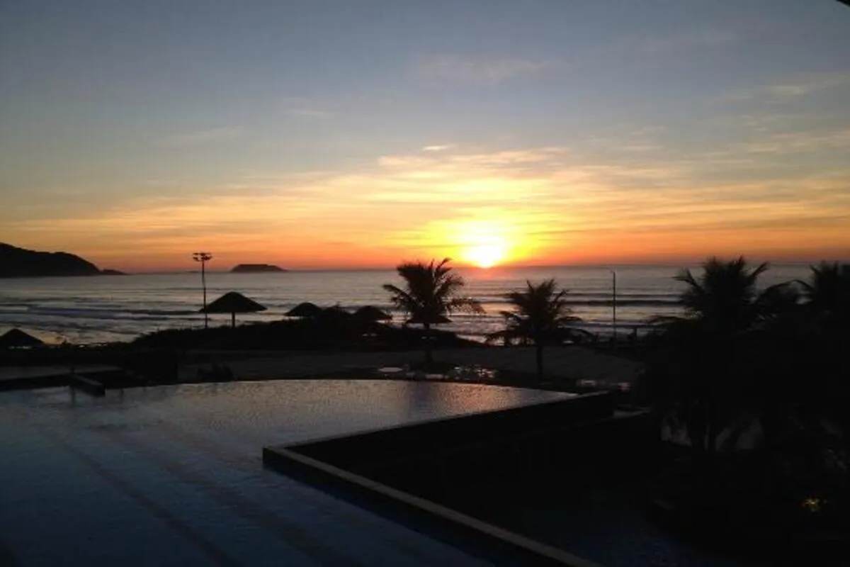 Pôr do sol na praia com o sol se pondo no horizonte, palmeiras e quiosques à beira-mar, criando um cenário tropical e relaxante.
