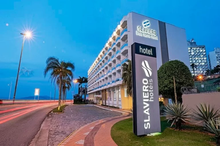 A fachada noturna do hotel Slaviero, com o prédio iluminado, o letreiro moderno com o logo da marca na frente, e rastros de luz de carros em uma rua próxima.