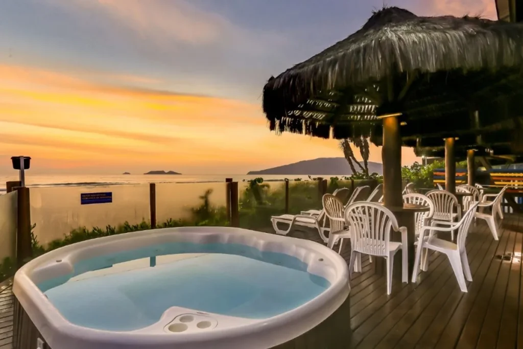 Área externa de hotel em Florianópolis com jacuzzi, cadeiras e vista para o mar ao pôr do sol.
