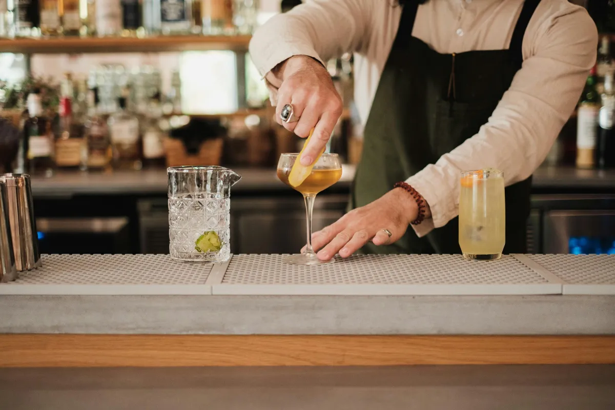 Barman preparando um drink no balcão de um bar, com garrafas e prateleiras ao fundo.