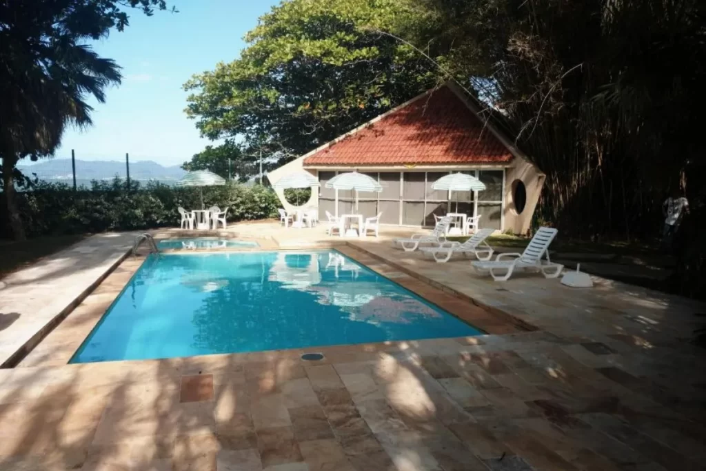 Piscina ao ar livre com espreguiçadeiras e casa de apoio ao fundo, cercada por árvores.