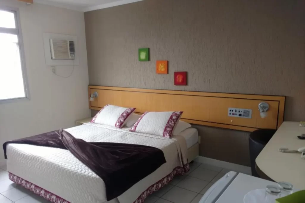Quarto de hotel com cama de casal arrumada, bancada lateral e decoração simples nas paredes.