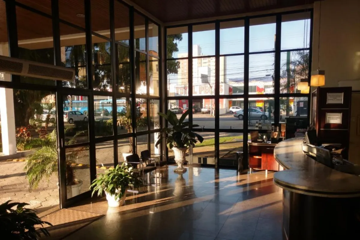 Lobby de hotel com paredes de vidro, poltronas, plantas decorativas e vista urbana ao fundo.
