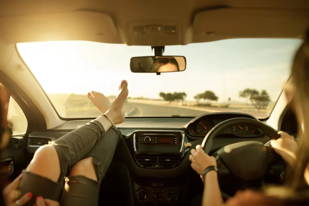 Vista interna de carro em viagem, com motorista ao volante e passageiro com os pés apoiados no painel.