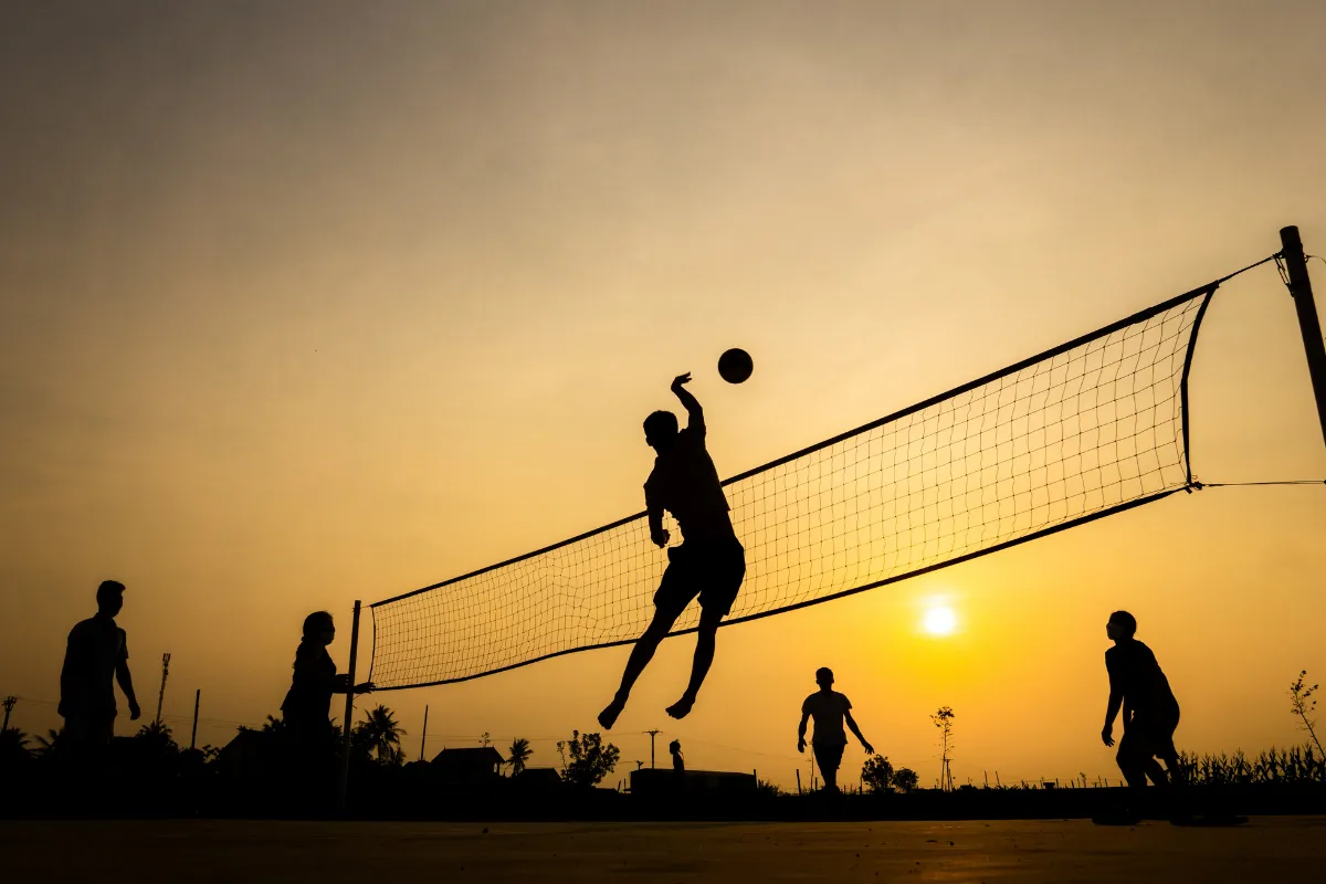 Jogadores praticando vôlei de praia ao pôr do sol, com silhuetas em destaque e rede montada na areia.