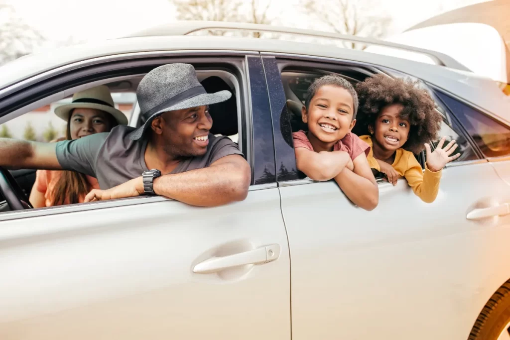 Família sorrindo pela janela de um carro durante viagem de carro em dia ensolarado.