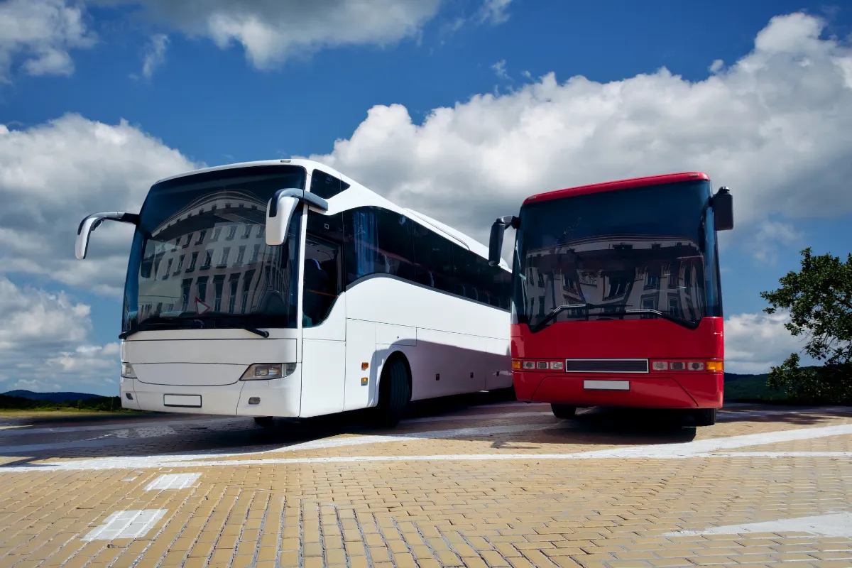 Dois ônibus, um branco e outro vermelho, estacionados em área pavimentada com céu azul e nuvens em Florianópolis.