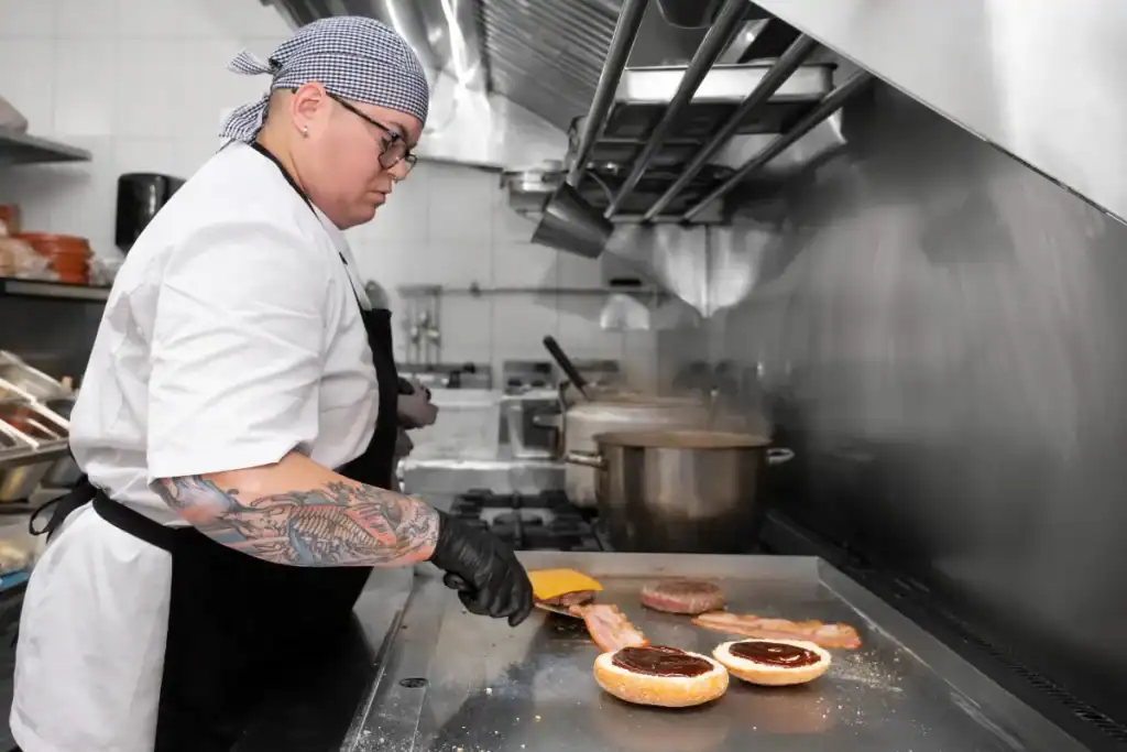 Hamburguerias em Florianópolis. Cozinheira preparando hambúrguer na chapa com bacon, pão e utensílios em cozinha profissional
