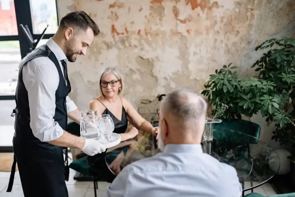 Garçom segurando bandeja com copos enquanto atende clientes sentados à mesa em restaurante.
