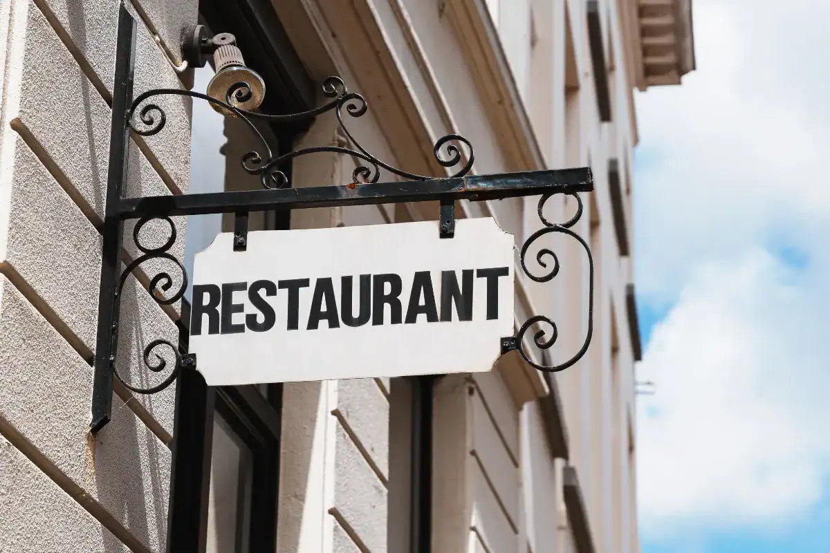 Placa com a palavra restaurant pendurada em suporte de ferro na fachada de um prédio.