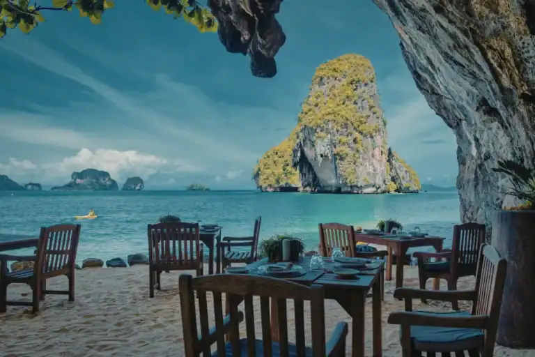 mesas de restaurante posicionadas na areia com vista direta para o mar e formações rochosas ao fundo