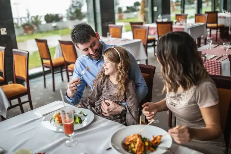 família com criança pequena sorrindo enquanto come em restaurante com mesas organizadas e ambiente iluminado