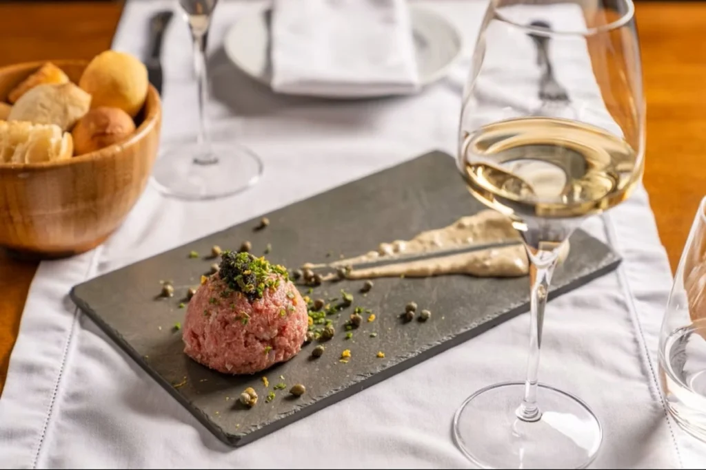 prato gourmet com steak tartare servido em tábua de pedra acompanhado de vinho branco e pão