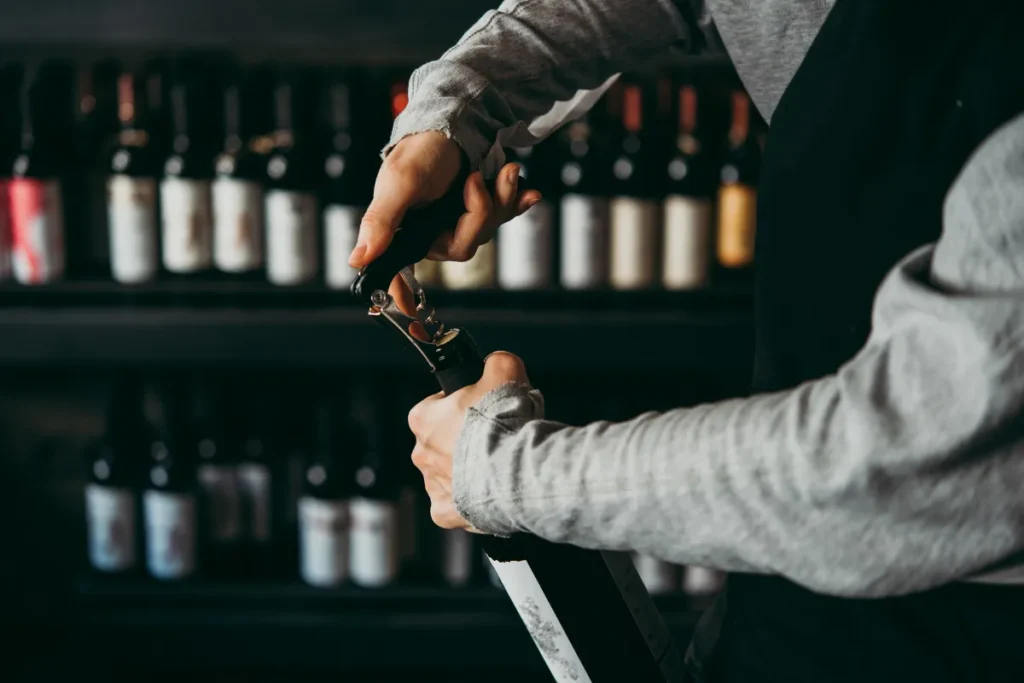 pessoa abrindo uma garrafa de vinho com saca-rolhas em frente a uma prateleira de bebidas