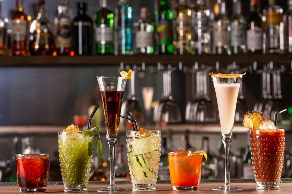 copos com diferentes coquetéis coloridos alinhados sobre o balcão de um bar com garrafas ao fundo