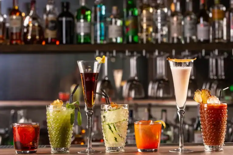 copos com diferentes coquetéis coloridos alinhados sobre o balcão de um bar com garrafas ao fundo
