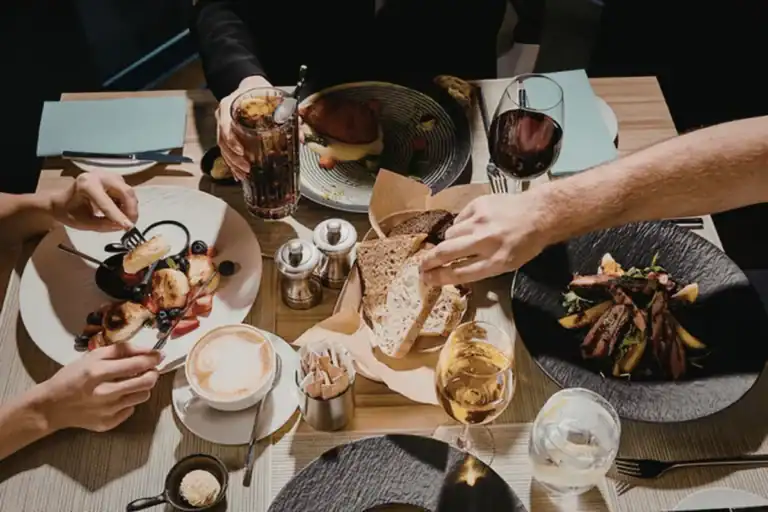 pessoas compartilhando pratos de brunch em uma mesa com pães, café, frutas e bebidas