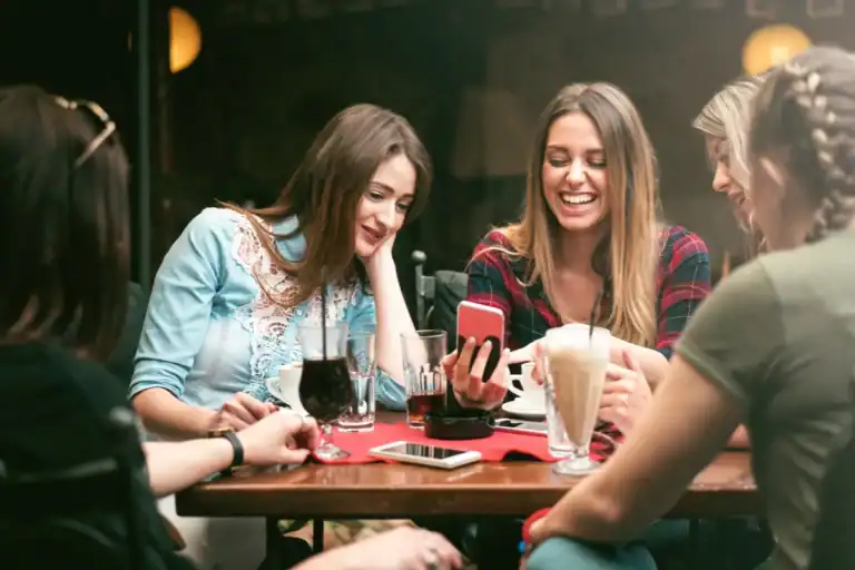 grupo de amigas sentadas à mesa em cafeteria sorrindo e olhando o celular com bebidas e cafés sobre a mesa