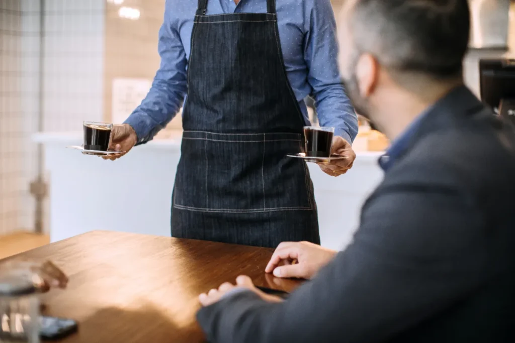 atendente de cafeteria servindo duas xícaras de café para cliente sentado à mesa
