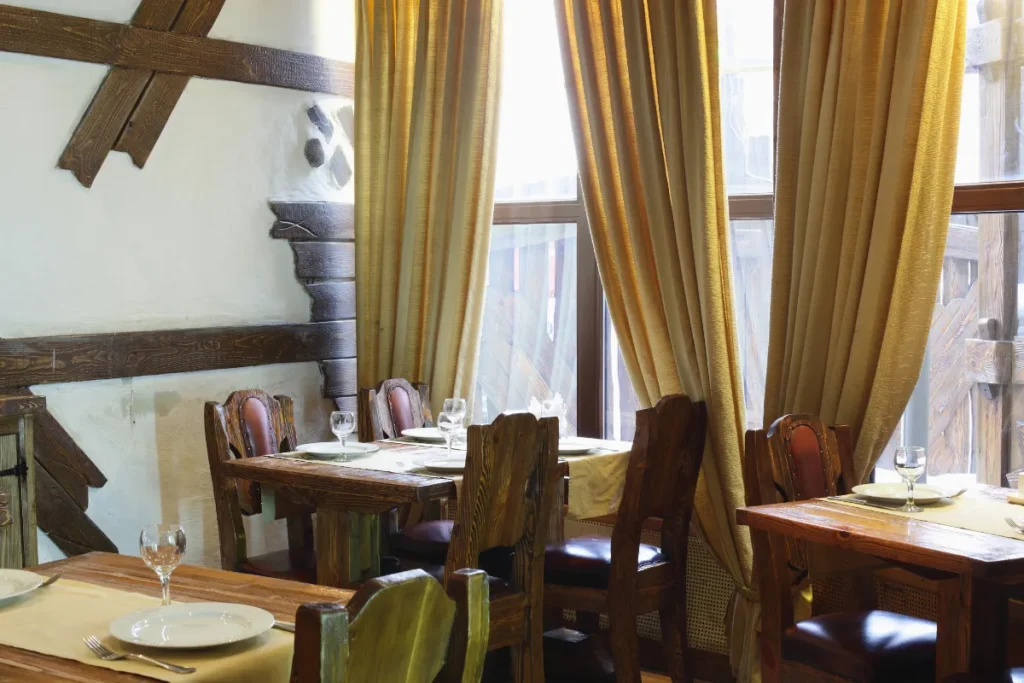 Onde comer em Jurerê. Interior de restaurante com mesas de madeira, cadeiras rústicas e cortinas grandes próximas a uma janela
