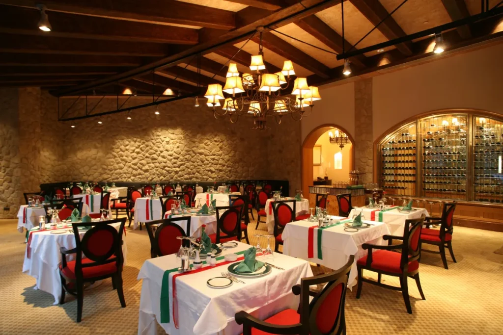 salão interno de restaurante com decoração rústica, mesas organizadas com toalhas e cadeiras vermelhas iluminadas por lustre