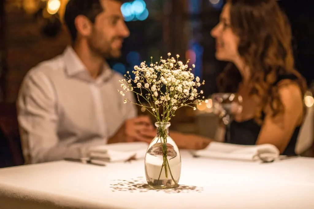 Mesa de restaurante com vaso de flores ao centro e casal ao fundo em ambiente noturno