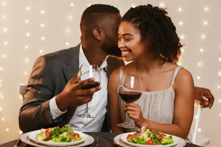 Casal sorrindo durante jantar com taças de vinho e pratos servidos em ambiente iluminado