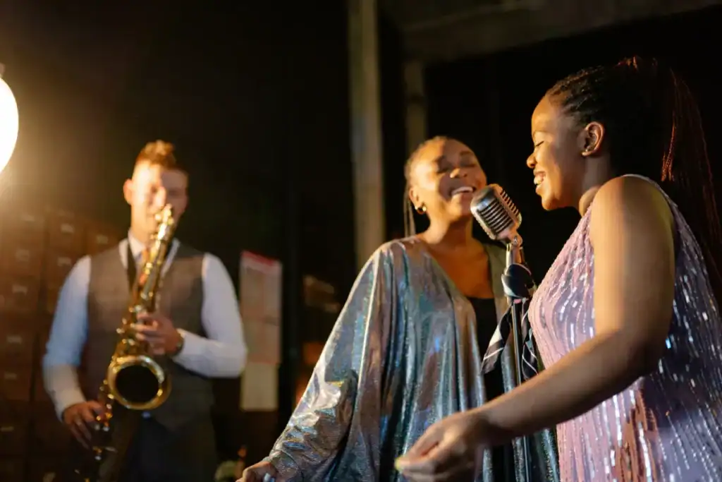 Cantoras se apresentando com microfone enquanto músico toca saxofone ao fundo em ambiente noturno