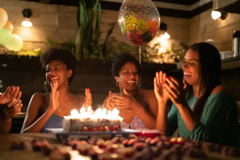 Grupo de mulheres sorrindo e batendo palmas ao redor de um bolo com velas acesas em uma comemoração de aniversário