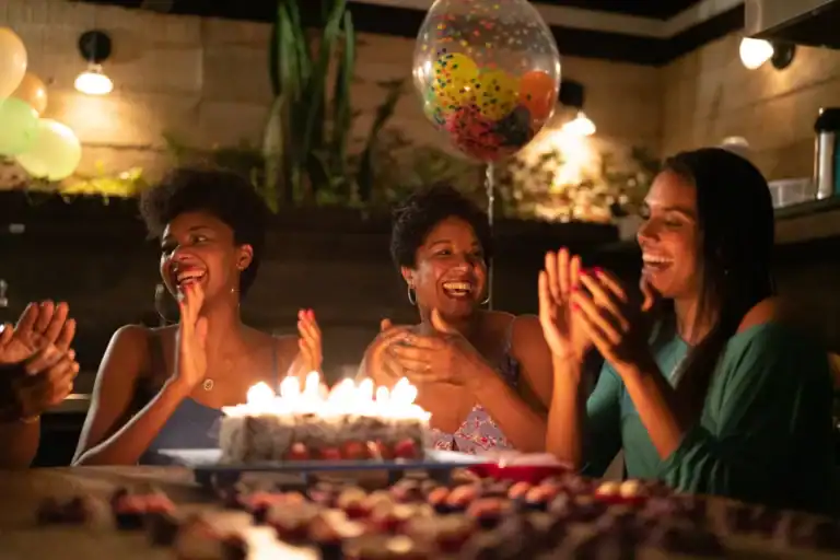 Grupo de mulheres sorrindo e batendo palmas ao redor de um bolo com velas acesas em uma comemoração de aniversário