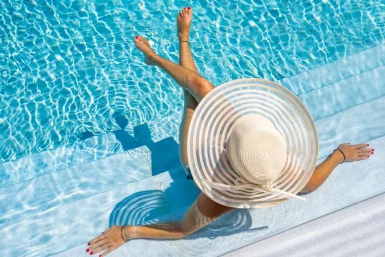 Pessoa relaxando na borda da piscina de resort com chapéu de praia em cenário tropical sofisticado.