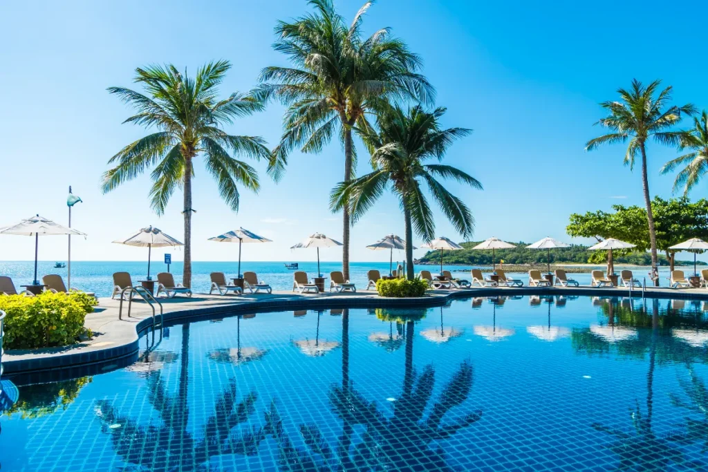 Piscina de resort à beira-mar com coqueiros, cadeiras de descanso e vista para o oceano em dia ensolarado.