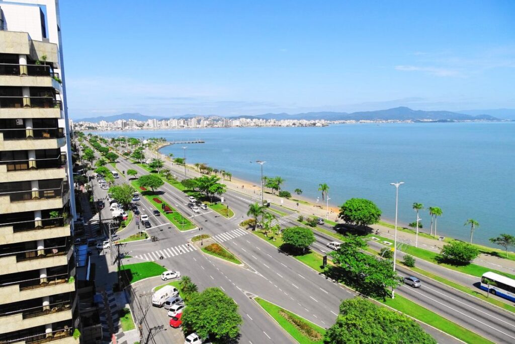 Vista aérea da Avenida Beira-Mar Norte em Florianópolis, com prédios, pista à beira-mar, árvores e baía azul ao fundo.