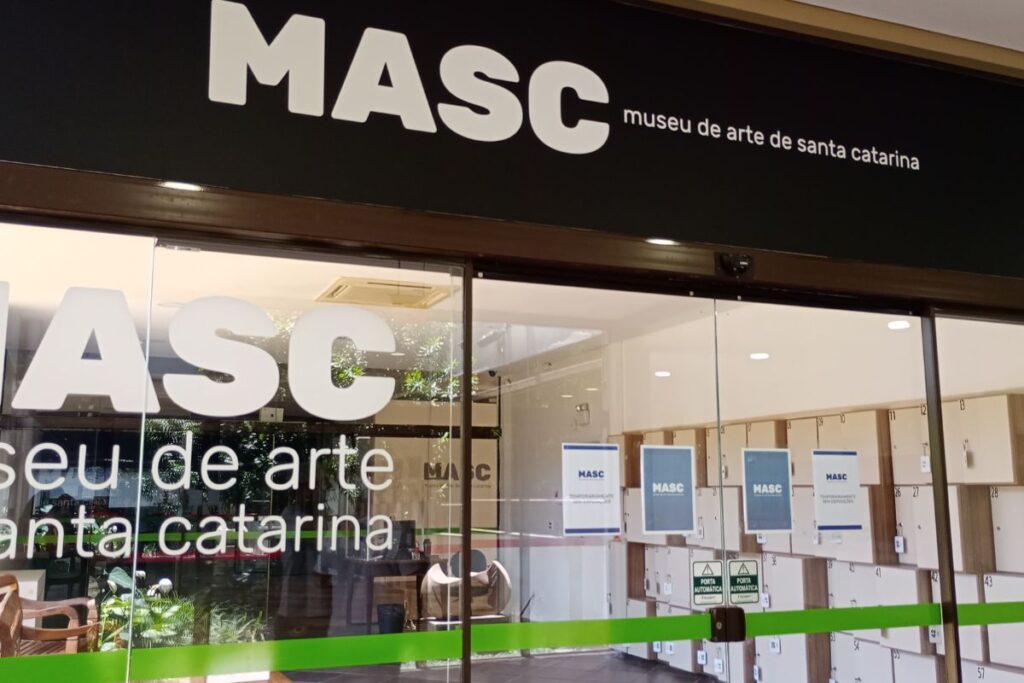 Entrada do Museu de Arte de Santa Catarina, o MASC, com fachada de vidro, identificação do museu e armários no interior.