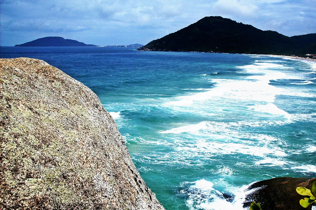 Vista da Praia Brava em Florianópolis com faixa de areia, ondas e ocupação urbana ao fundo